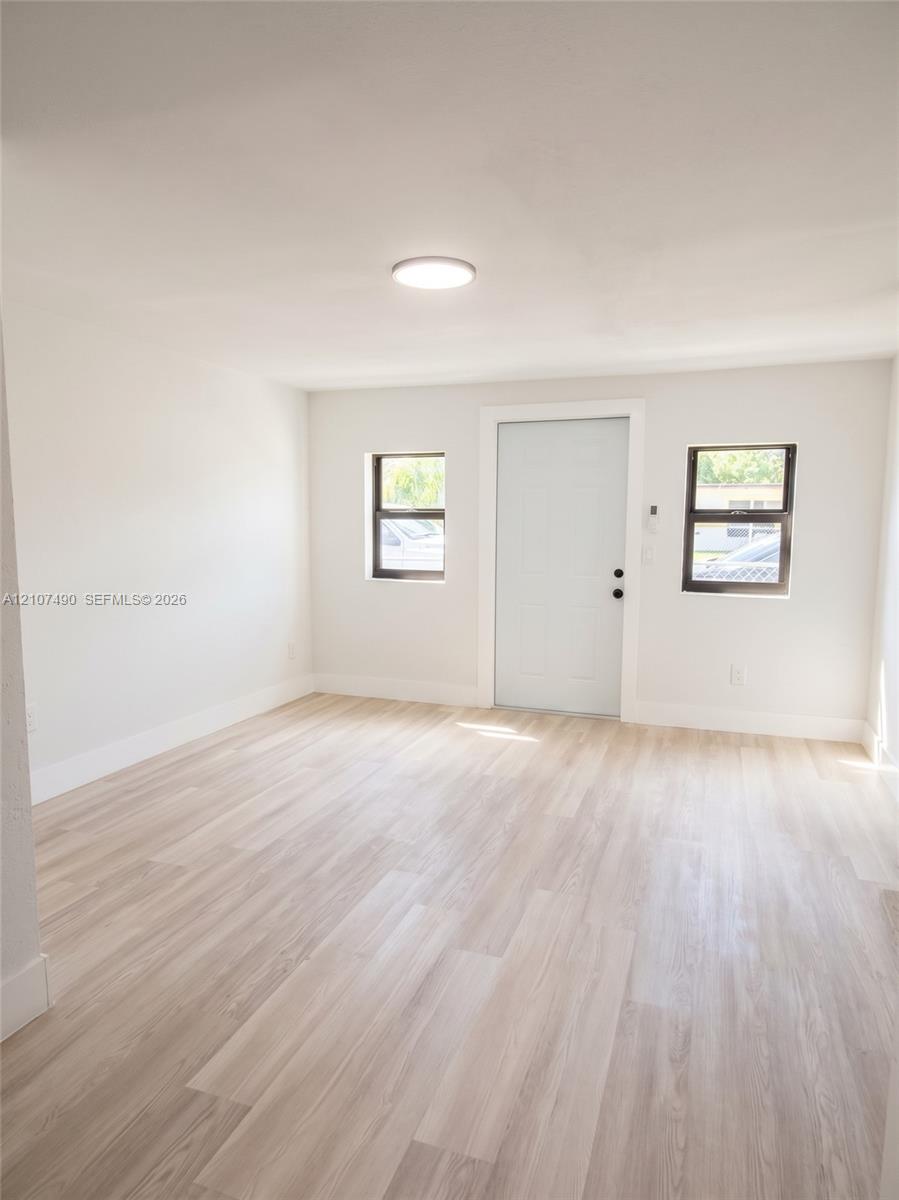 Spacious Interior space featuring light-toned wood-finish flooring, neutral wall paint, two windows with dark frames, and a solid-core entry door