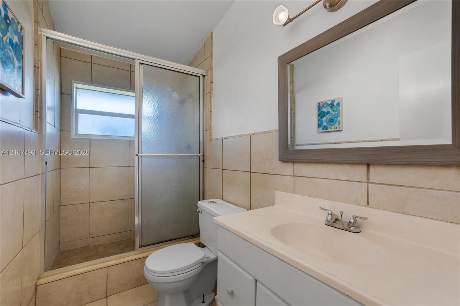 Bathroom featuring a vanity with an integrated basin, a framed wall mirror, a tiled shower enclosure with sliding glass doors, and light-toned wall tile