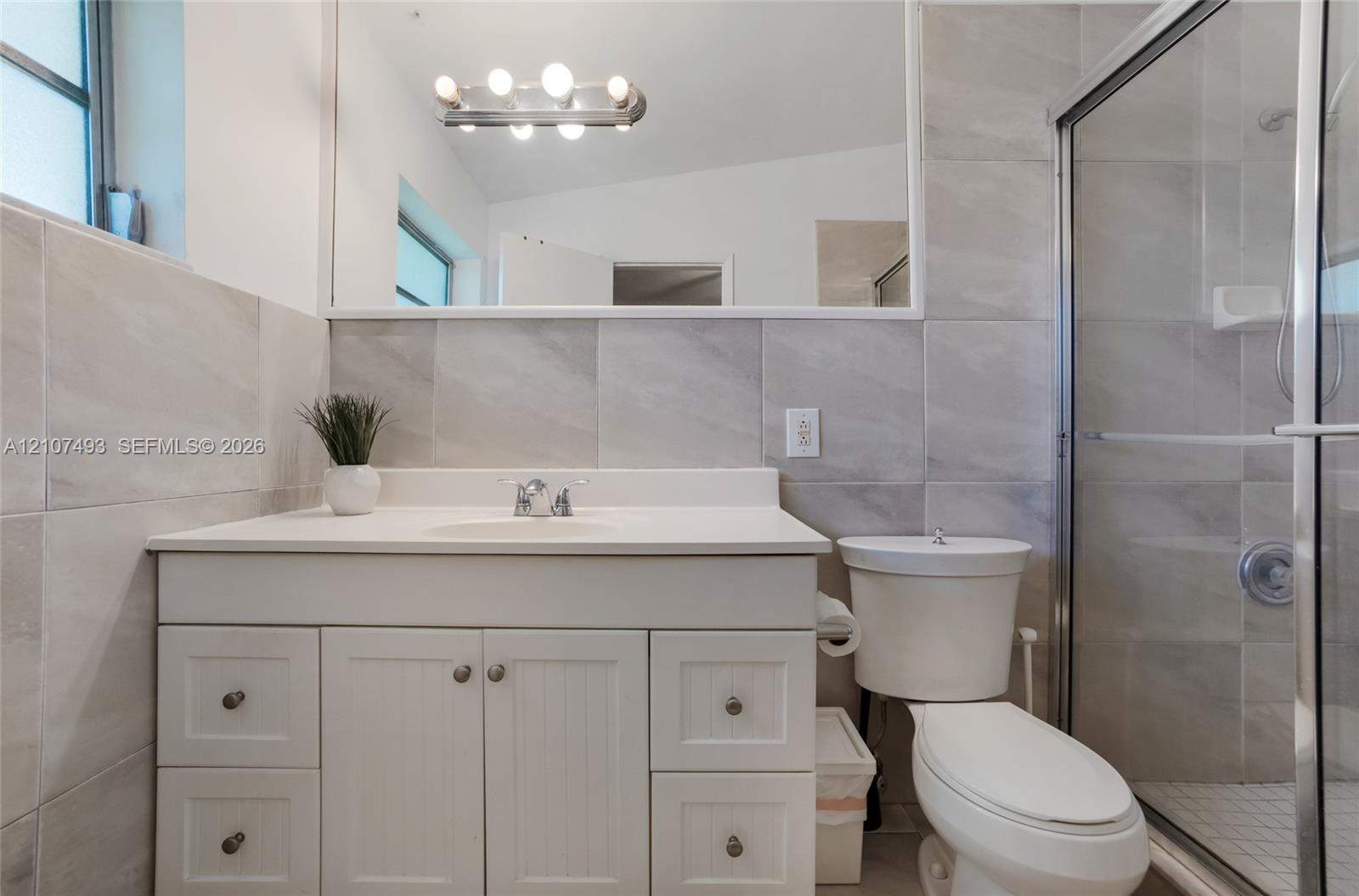 Bathroom featuring a white vanity cabinet, solid-surface countertop with integrated sink, large-format tile wall cladding, full-width mirror, and a glass-enclosed shower