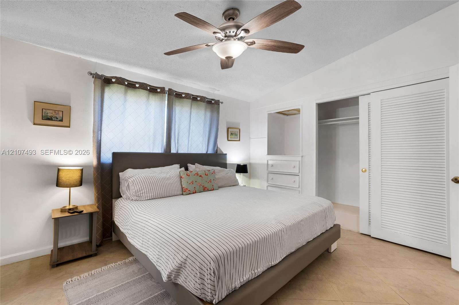 Neutral-toned tile flooring, ceiling fan with integrated lighting, built-in dresser with inset mirror, and sliding louvered closet doors