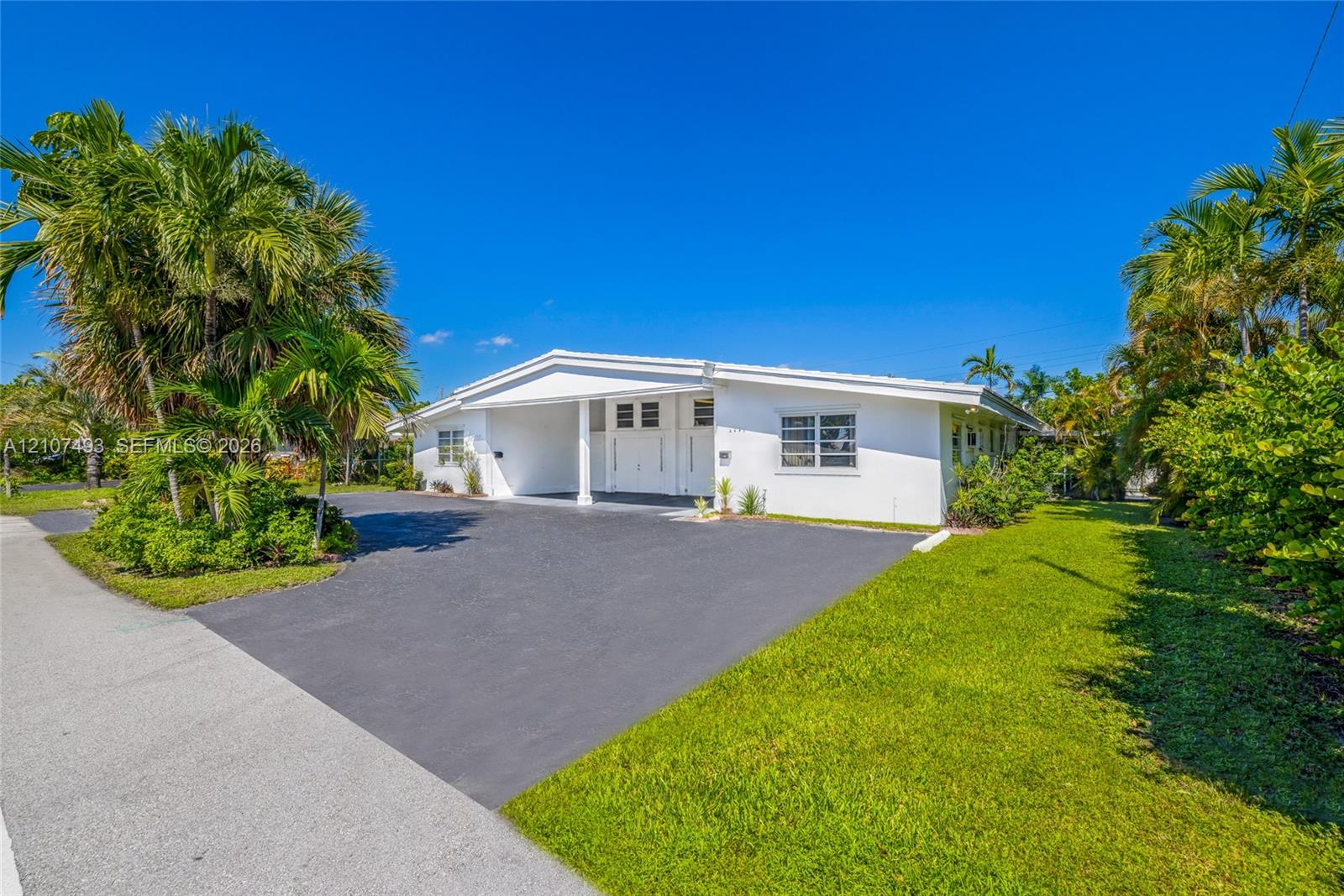 Single-story exterior featuring white stucco cladding, a paved driveway, a covered entrance, horizontal louvered windows, and mature palm trees