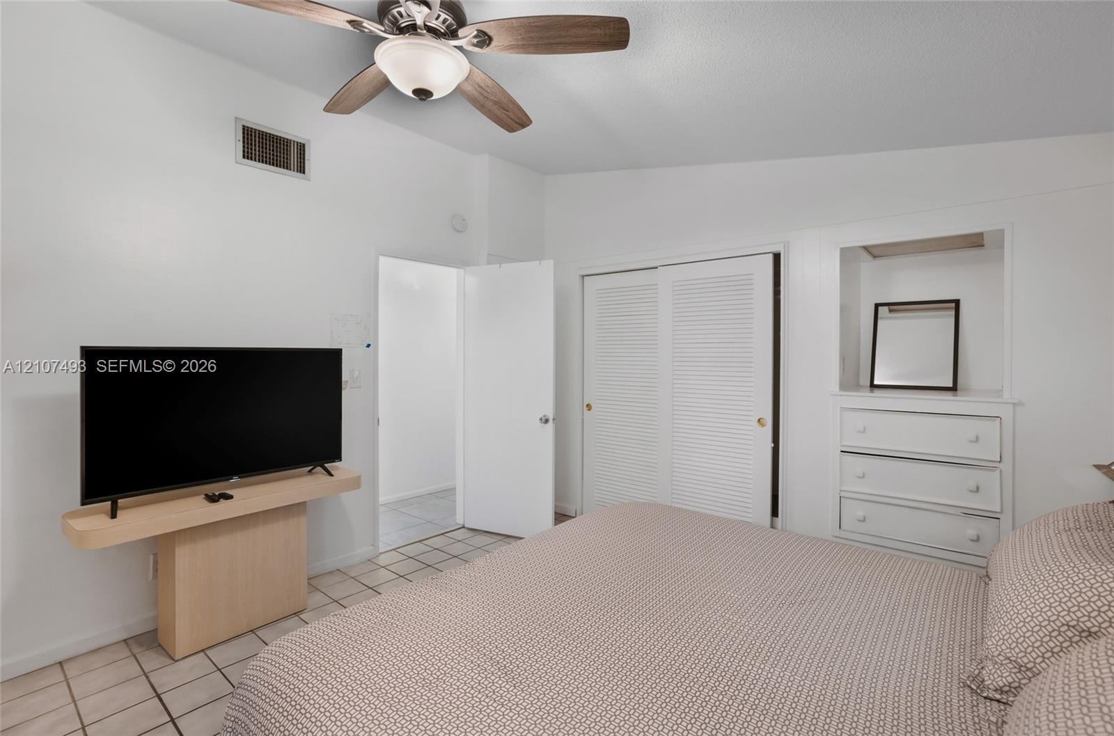 Room featuring vaulted ceilings, neutral ceramic tile flooring, ceiling fan with integrated lighting, built-in wooden shelving, and a closet with louvered doors