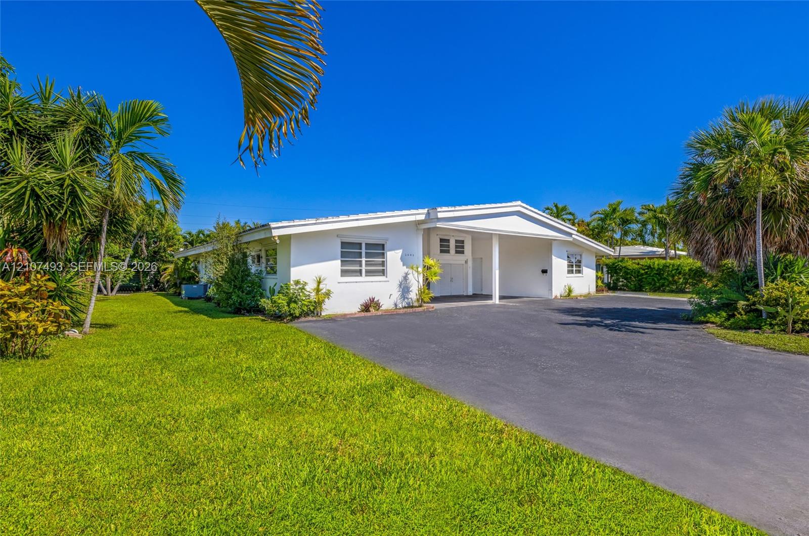 Single-story residence featuring white exterior cladding, a covered carport, and asphalt driveway, surrounded by mature palm trees and landscaped grounds