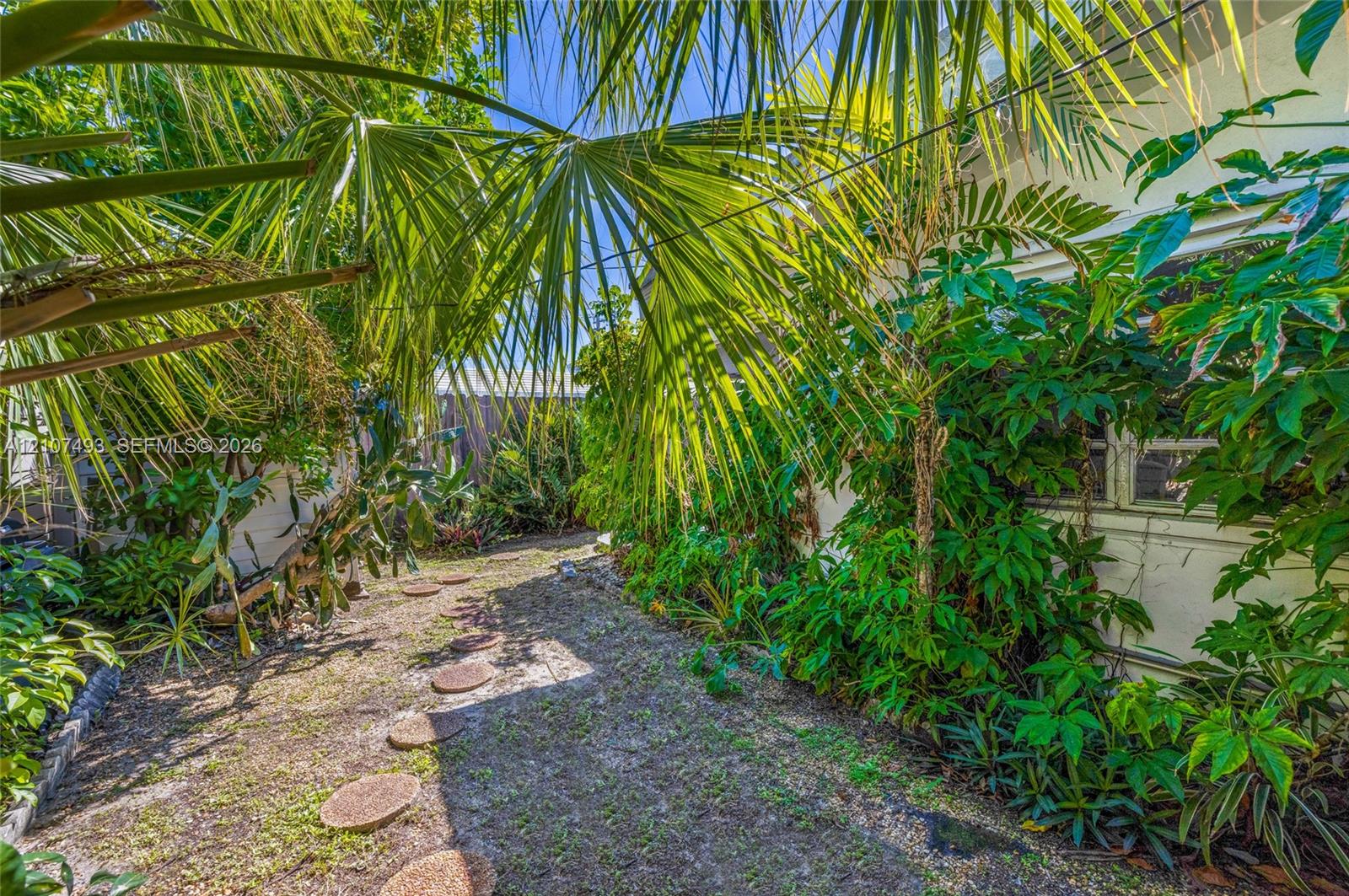 Exterior side yard featuring mature palm trees, dense tropical greenery, and a stone paver pathway