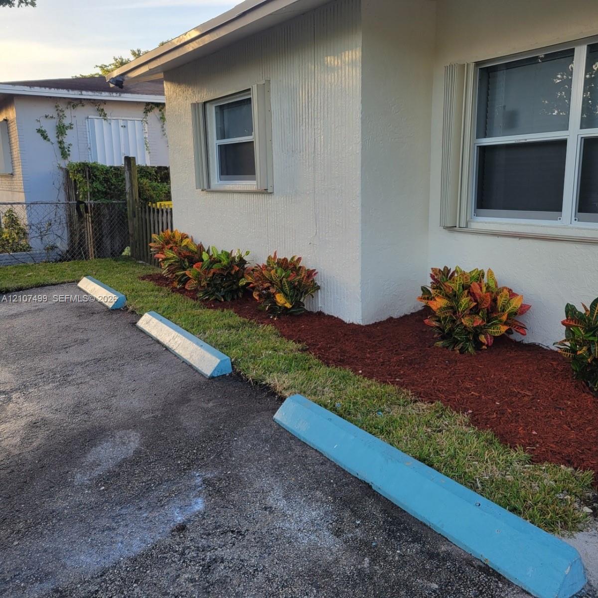 Exterior facade featuring stucco siding, multi-pane windows, paved parking area with concrete wheel stops, and landscaped garden beds with decorative mulch Exterior facade featuring stucco siding, multi-pane windows, paved parking area with concrete wheel stops, and landscaped garden beds with decorative mulch