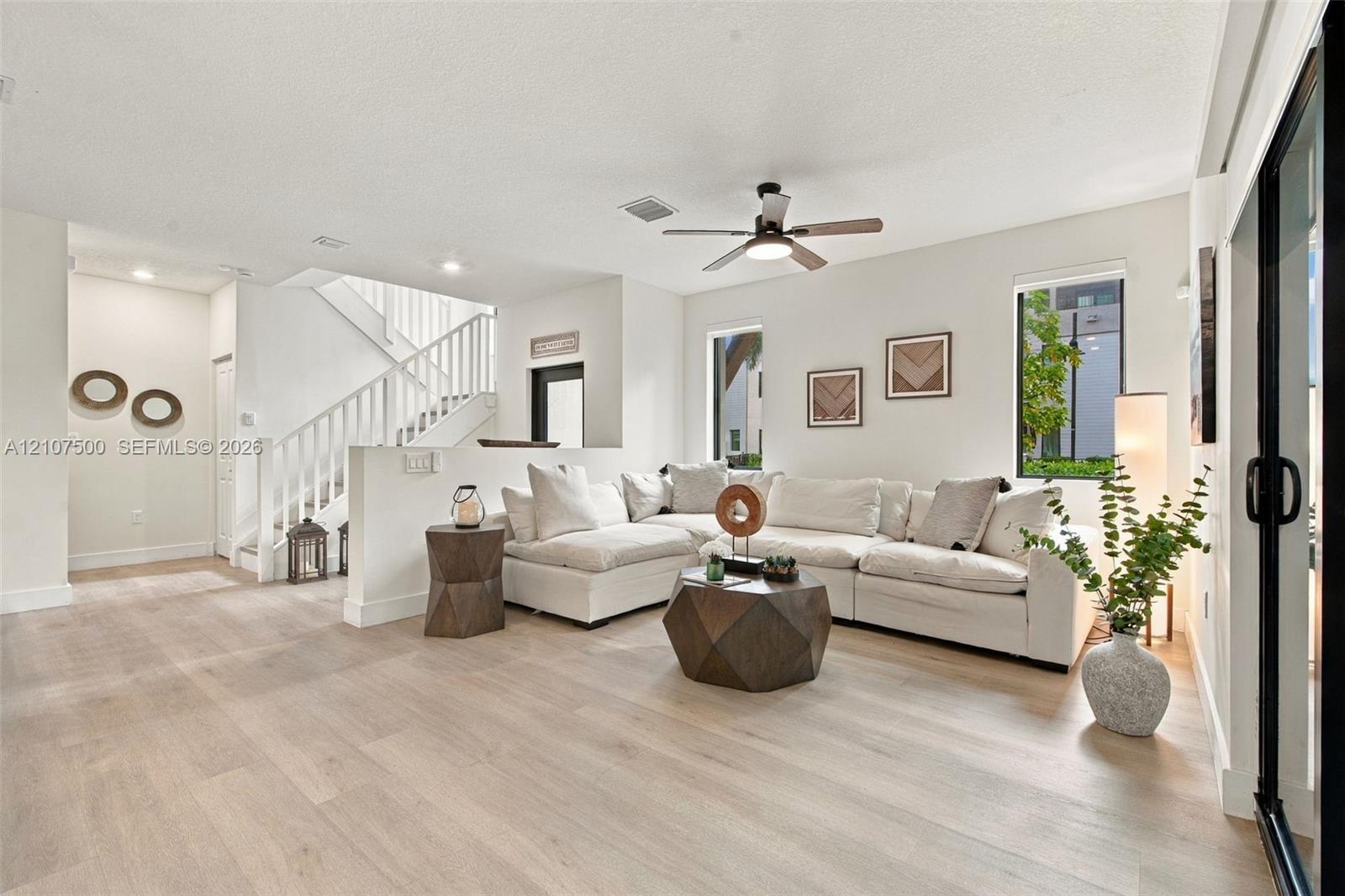Open-concept interior featuring wood-finish flooring, a white staircase with a vertical balustrade, recessed lighting, a ceiling fan with integrated lighting, and glass sliding doors