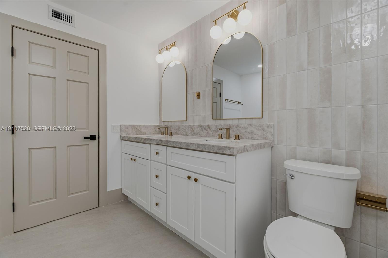 Dual-sink vanity with white cabinetry and stone-finish countertops, glass-enclosed shower with gold-tone hardware, pebble-stone shower flooring, and arched wall mirrors with integrated lighting