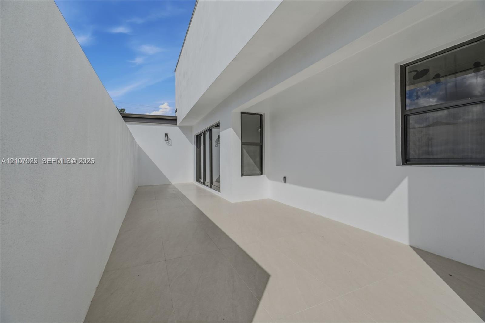 Modern exterior patio featuring large-format porcelain tile flooring, expansive floor-to-ceiling sliding glass panels, smooth white stucco finish, and a private enclosed yard