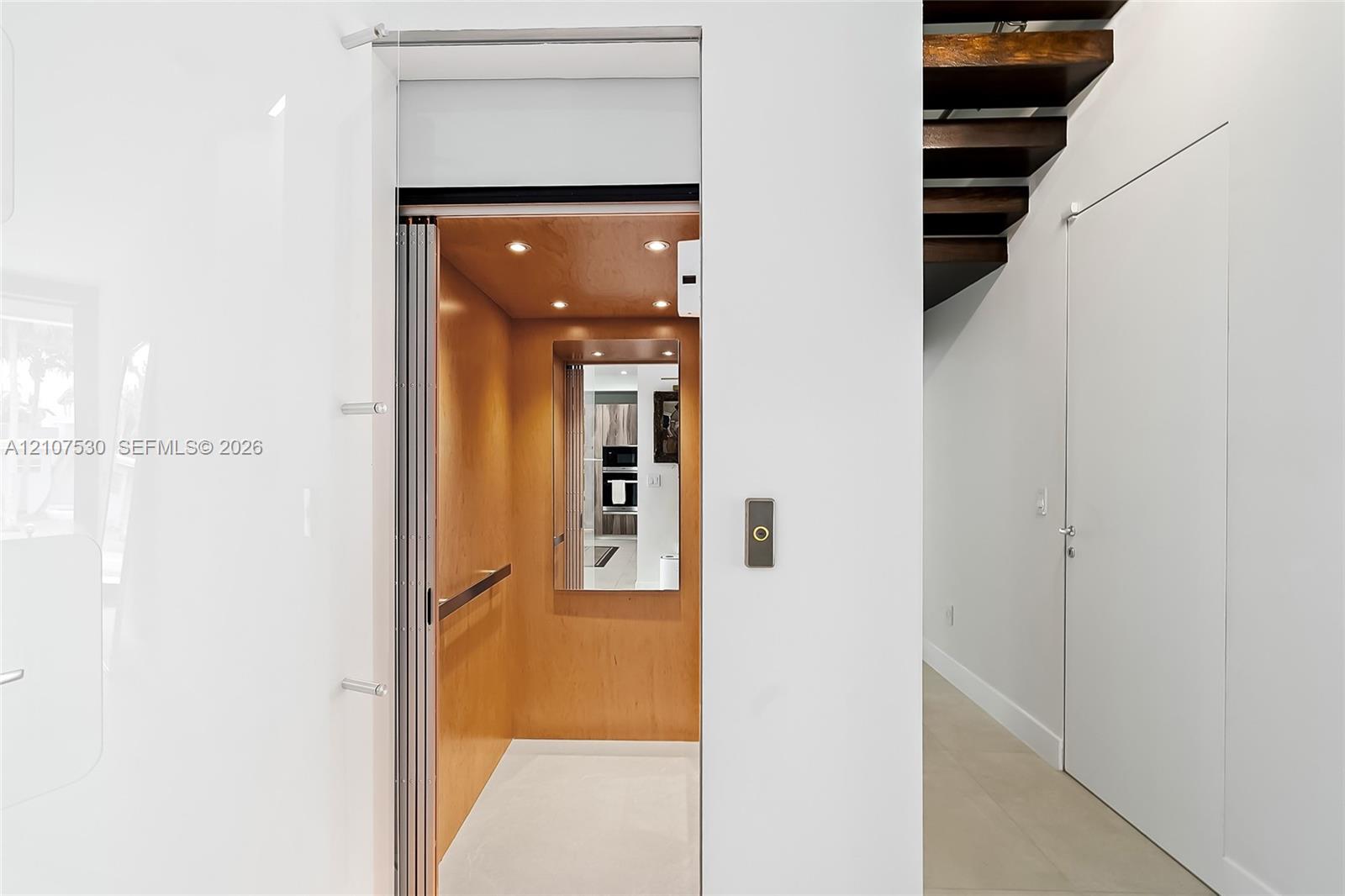 Residential elevator featuring a wood-paneled interior, recessed lighting, and a mirrored wall, complemented by an open-riser staircase with dark wood treads and neutral stone flooring