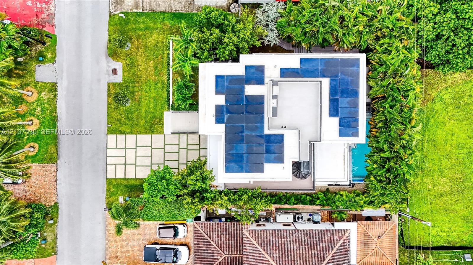 Aerial perspective featuring a flat-roof residence equipped with a solar panel array, paved concrete grid driveway, perimeter landscaping, and a ground-level swimming pool