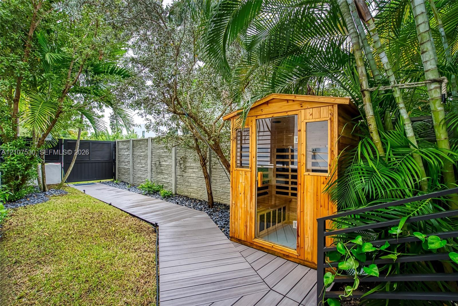 Wood-finished exterior sauna featuring a glass door and paneling, surrounded by composite decking, a textured masonry perimeter wall, and mature landscaping
