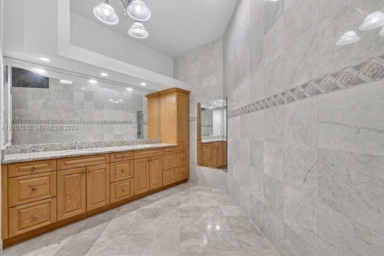 Vanity featuring light-toned wood cabinetry, stone countertop, dual undermount sinks, and full-width mirror, complemented by floor-to-ceiling wall tiling with decorative tile inlay and large-format tile flooring
