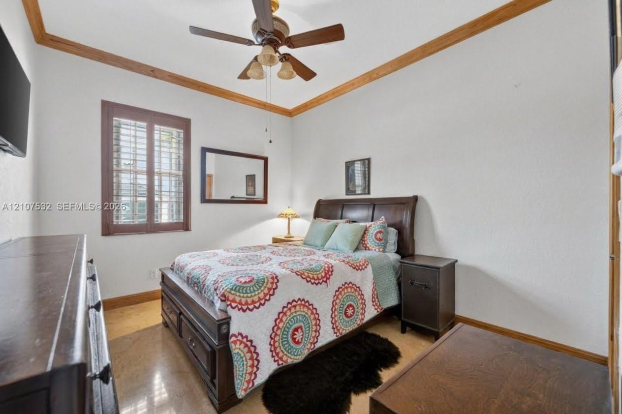 Room featuring wood-finish ceiling crown molding, a central ceiling fan with integrated lighting, polished stone flooring, and window shutter accents Room featuring wood-finish ceiling crown molding, a central ceiling fan with integrated lighting, polished stone flooring, and window shutter accents