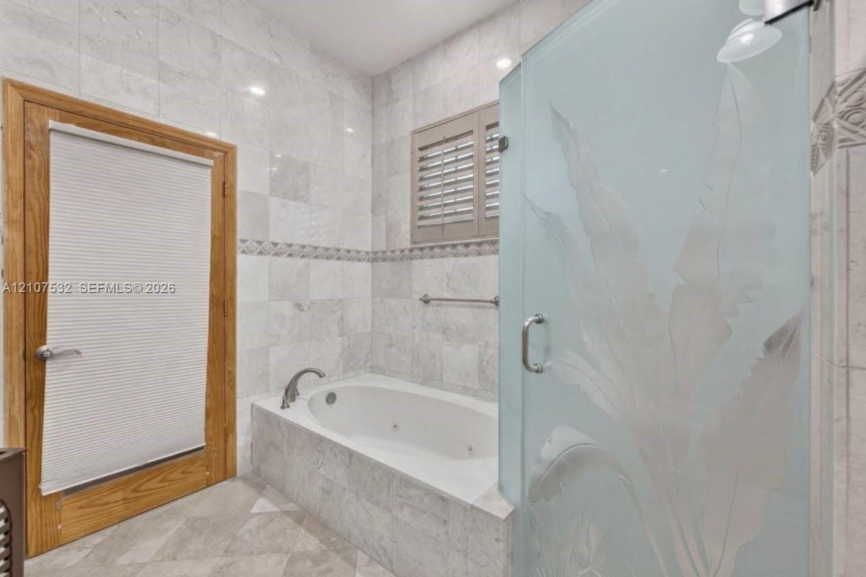 Jetted soaking tub set in a tile-surrounded deck, walk-in shower with decorative etched-glass enclosure, natural stone wall tiling with an accent border, and wood-finish door trim