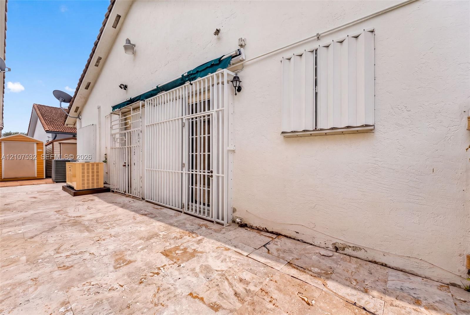 Exterior area featuring a neutral-toned stone-tile patio, white stucco siding, metal security shutters, and an attached retractable awning