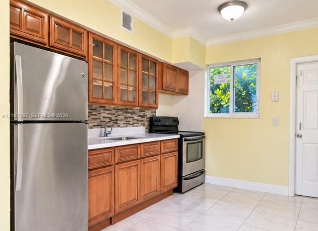 Kitchen featuring wood cabinetry with glass-front uppers, stainless steel appliances, multi-toned tile backsplash, solid-surface countertops, and tile flooring Kitchen featuring wood cabinetry with glass-front uppers, stainless steel appliances, multi-toned tile backsplash, solid-surface countertops, and tile flooring
