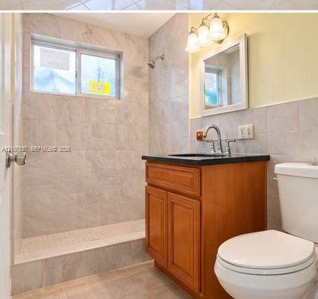 Bathroom featuring a wood-finish vanity with a dark stone-top surface, wall-mounted three-light fixture, framed mirror, and tile-surround walk-in shower Bathroom featuring a wood-finish vanity with a dark stone-top surface, wall-mounted three-light fixture, framed mirror, and tile-surround walk-in shower