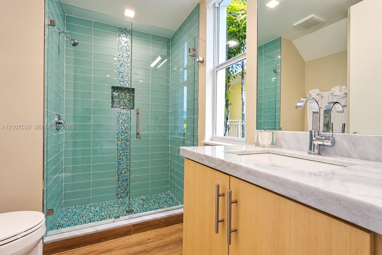 Walk-in shower with horizontal tile surround, mosaic accent strip, and glass enclosure, paired with a vanity featuring a light stone countertop, an integrated rectangular sink, and chrome fixtures