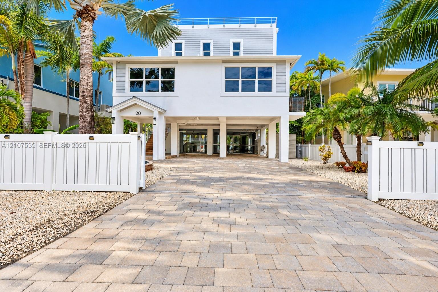Three-story elevated residence featuring a paver driveway, covered ground-level parking, multi-pane windows, and a white vinyl perimeter fence