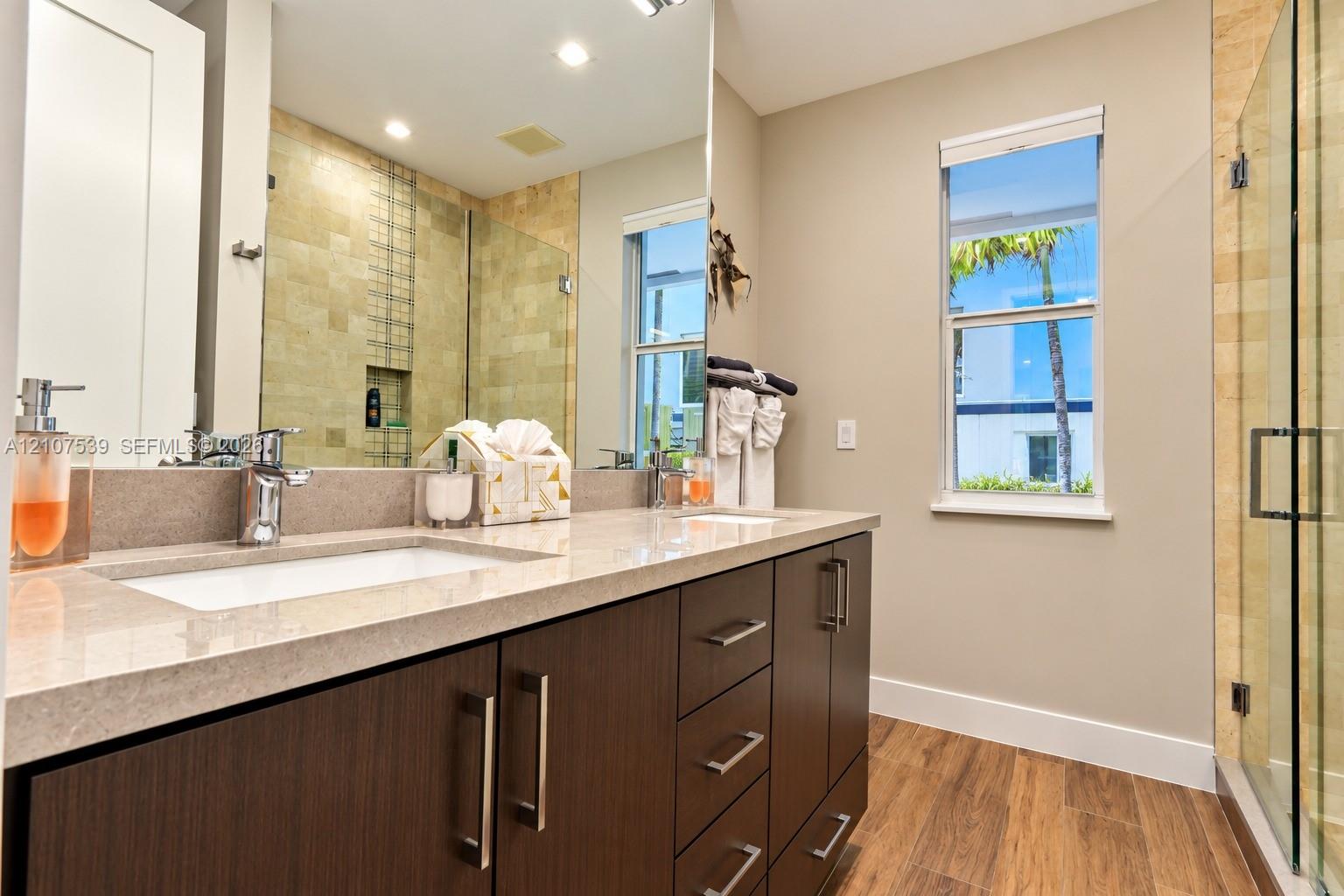 Dual-sink vanity with composite countertop, cabinetry with metallic hardware, wood-finish flooring, and a glass-enclosed shower with tiled wall accents