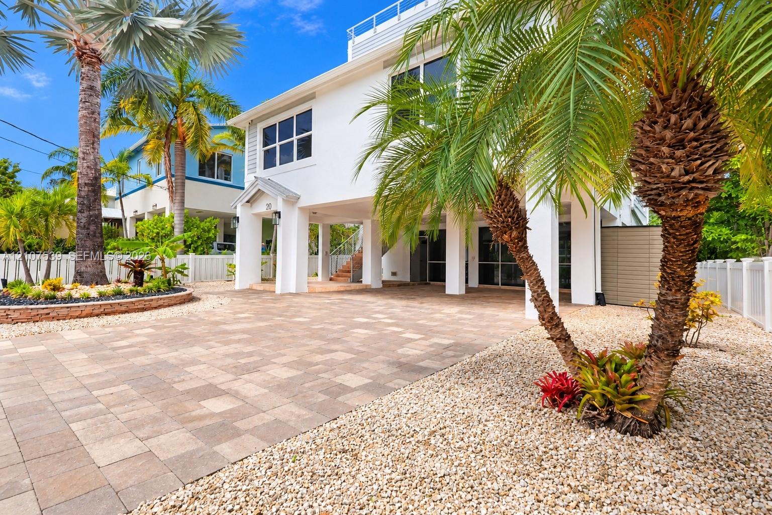 Two-story elevated residence featuring a paver driveway, covered ground-level parking, white siding, and landscaped exterior grounds with palm trees