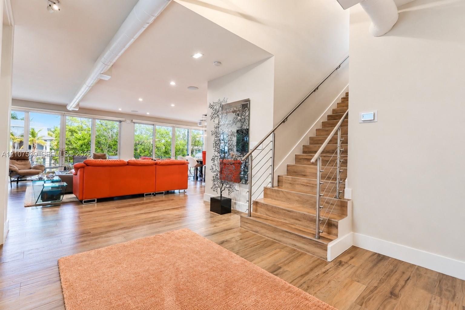 Open floor plan featuring wood-finish flooring, floor-to-ceiling windows, exposed industrial ductwork, recessed lighting, and a staircase with cable railings and wood-finish treads