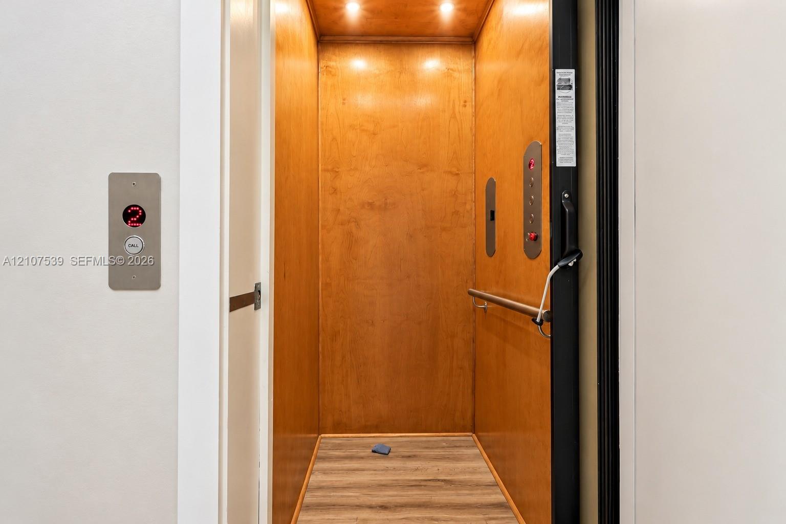 Private elevator featuring wood-paneled interior walls, integrated ceiling lighting, horizontal metal handrail, and wood-finish flooring