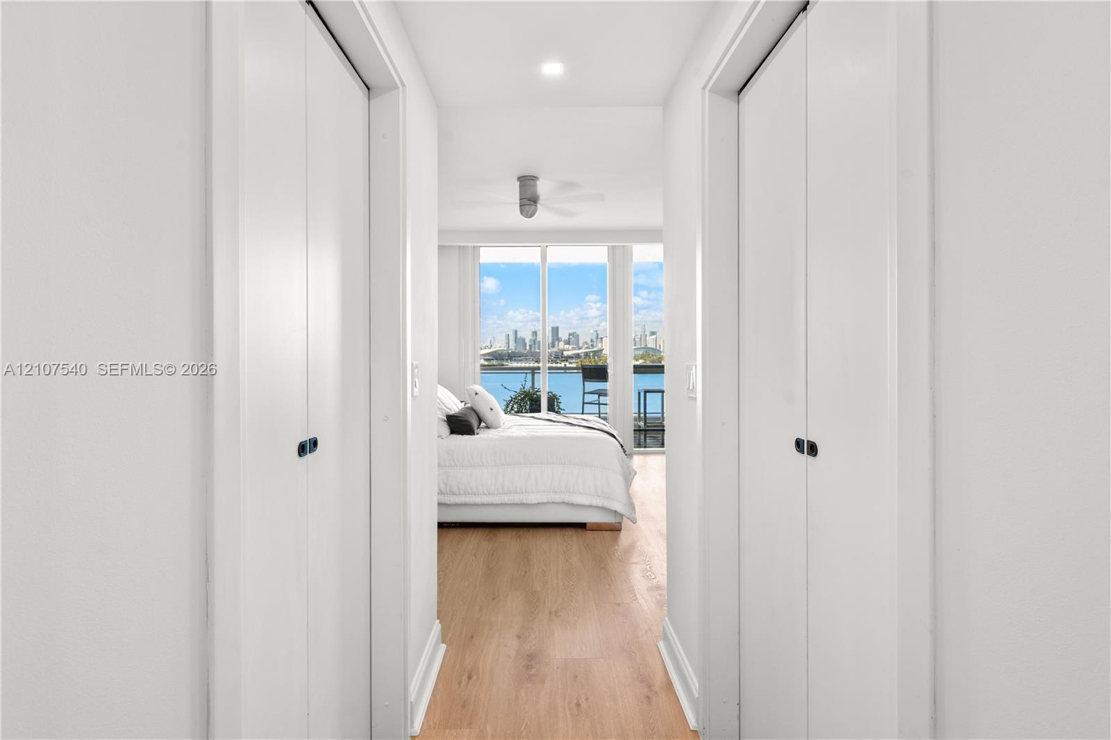 Master bedroom hallway with dual bifold closet doors, wood-finish flooring, recessed LED lighting, and floor-to-ceiling windows with views of the downtown Miami skyine + Biscayne Bay