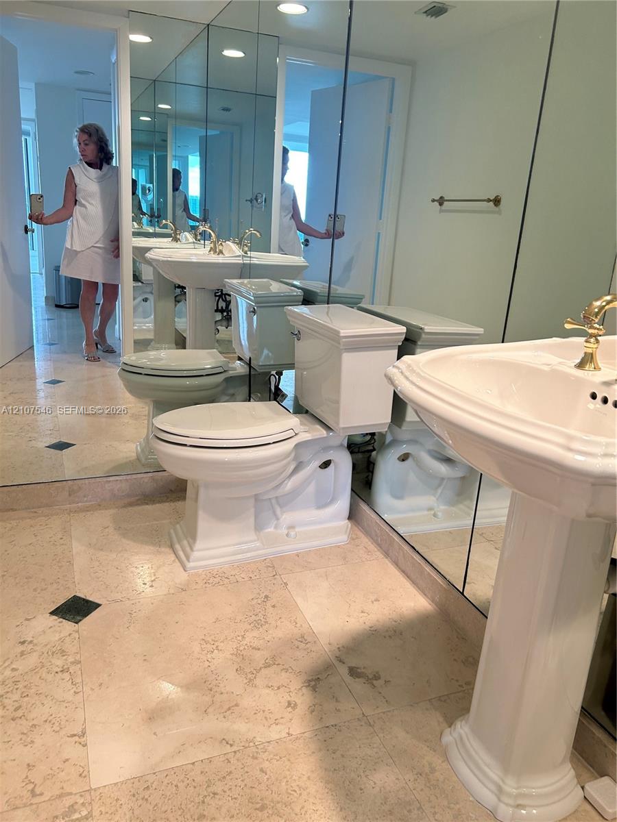 Bathroom featuring large-format light stone flooring with inlay accents, a pedestal sink with gold-tone hardware, and floor-to-ceiling wall mirrors