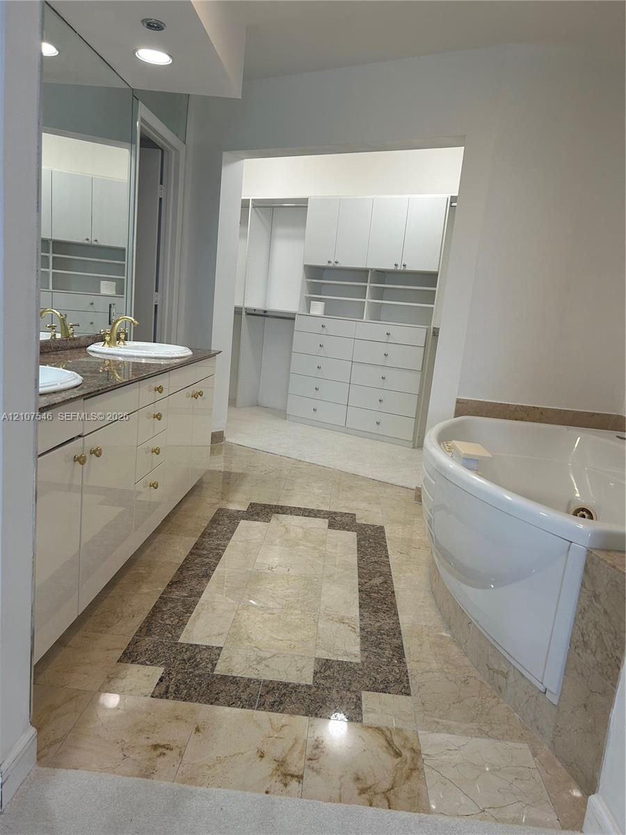 Dual-sink vanity with stone countertop and gold-tone fixtures, expansive wall mirror, jetted soaking tub with stone surround, and custom built-in wardrobe cabinetry with integrated drawer storage