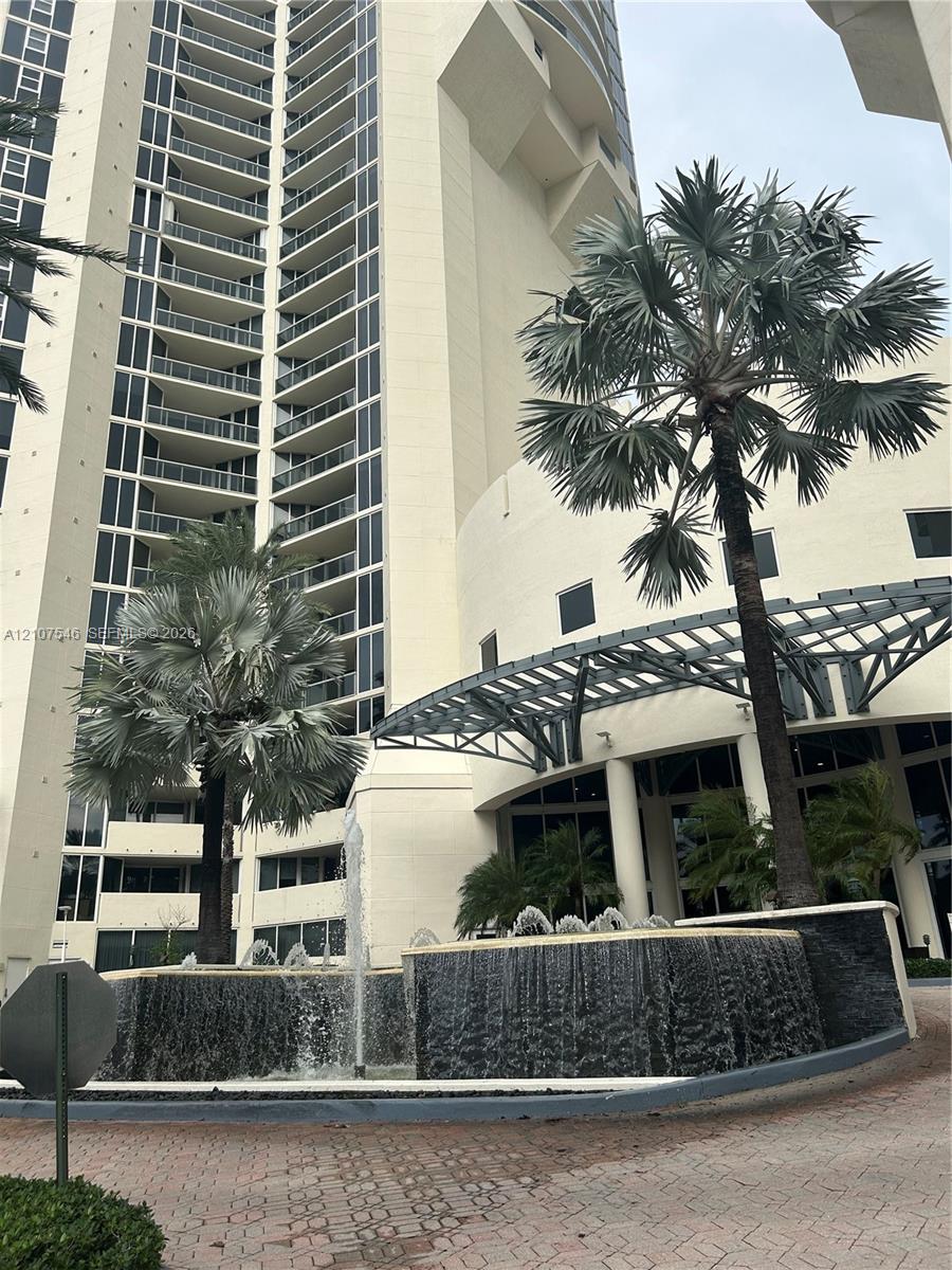 High-rise exterior featuring curved glass balconies, a circular fountain with tiered stone masonry, a metal-framed porte-cochere, and mature palm landscaping High-rise exterior featuring curved glass balconies, a circular fountain with tiered stone masonry, a metal-framed porte-cochere, and mature palm landscaping