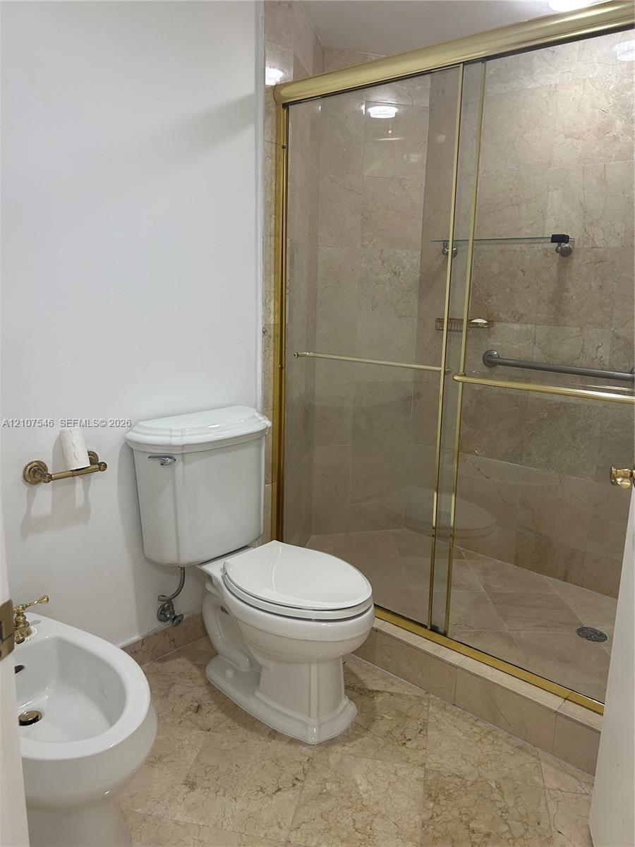 Bathroom featuring stone tile flooring and shower surround, glass-enclosed shower with metal framing, wall-mounted bidet, and white porcelain toilet