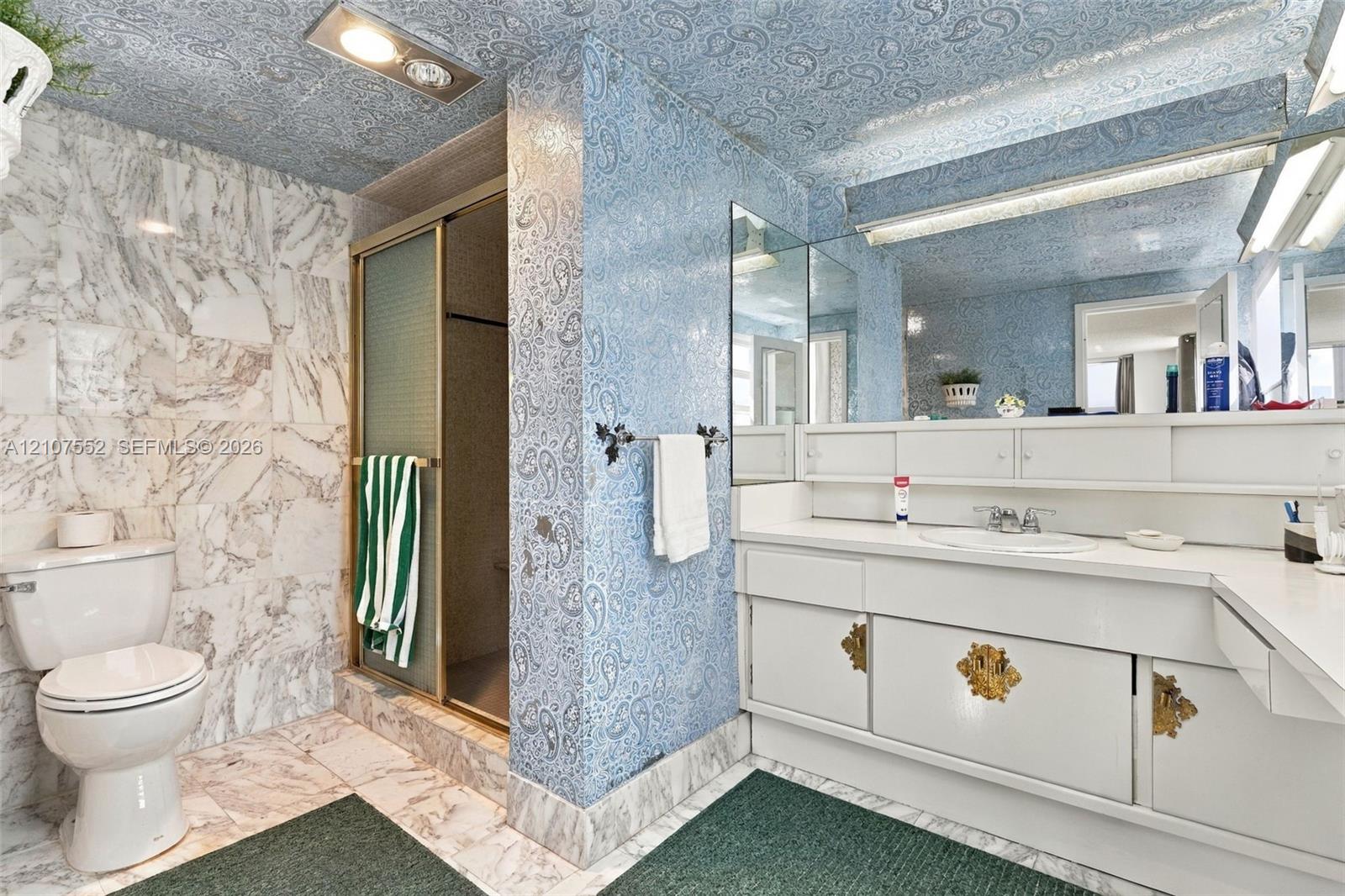 Bathroom featuring natural stone wall and floor tiling, walk-in shower with metal-framed glass enclosure, white vanity cabinetry, integrated full-width wall mirror, and patterned wall and ceiling covering