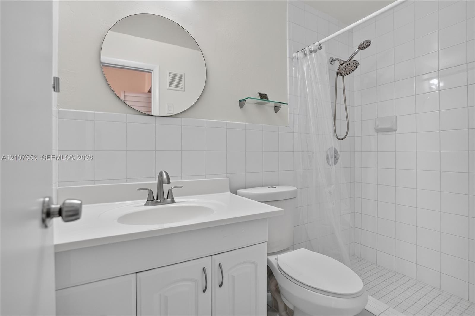 Bathroom featuring a white vanity cabinet with an integrated basin, circular wall-mounted mirror, glass shelf, and a walk-in shower enclosure with white wall tile
