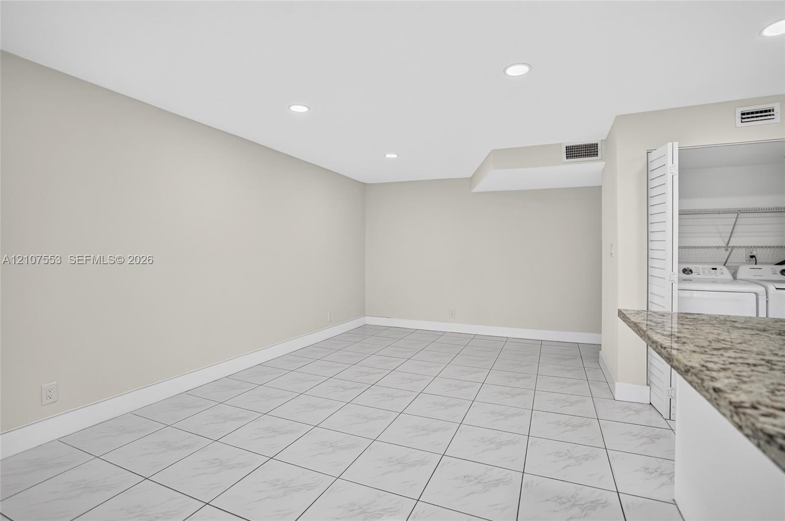 Neutral-toned interior space featuring light-colored tile flooring, recessed lighting, and a built-in stone-top counter with an integrated utility area behind louvered doors