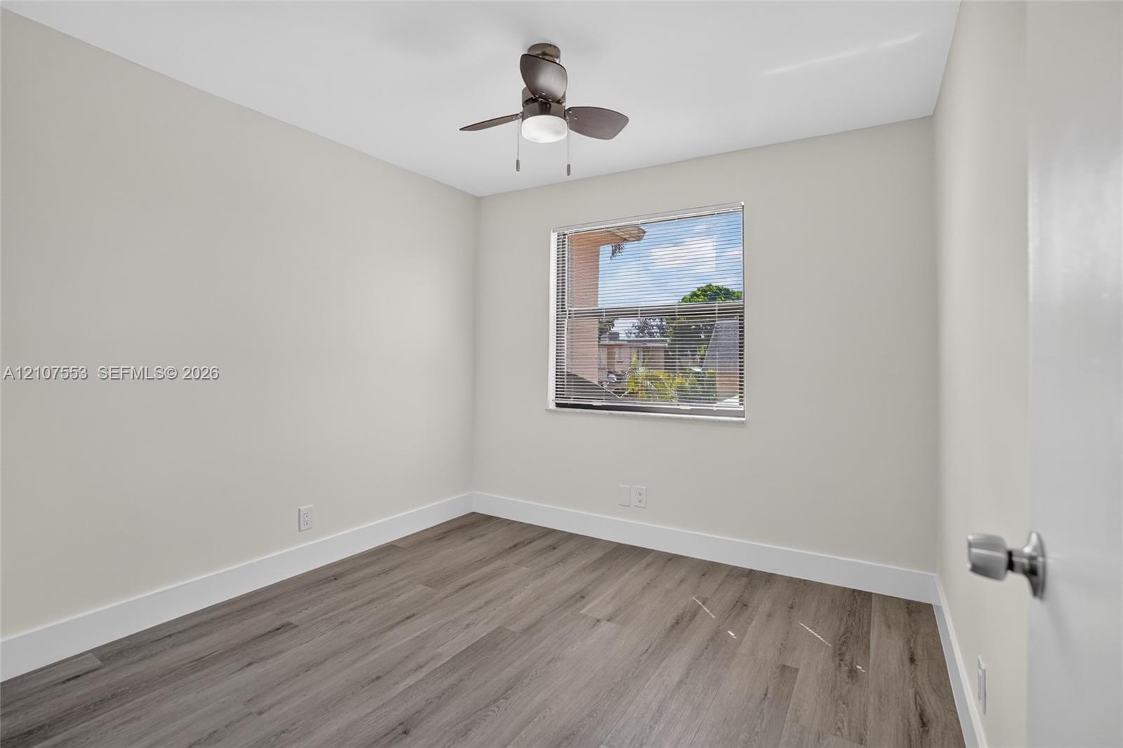 Neutral-toned interior room featuring wood-finish flooring, a ceiling fan with integrated light fixture, and a window with horizontal blinds