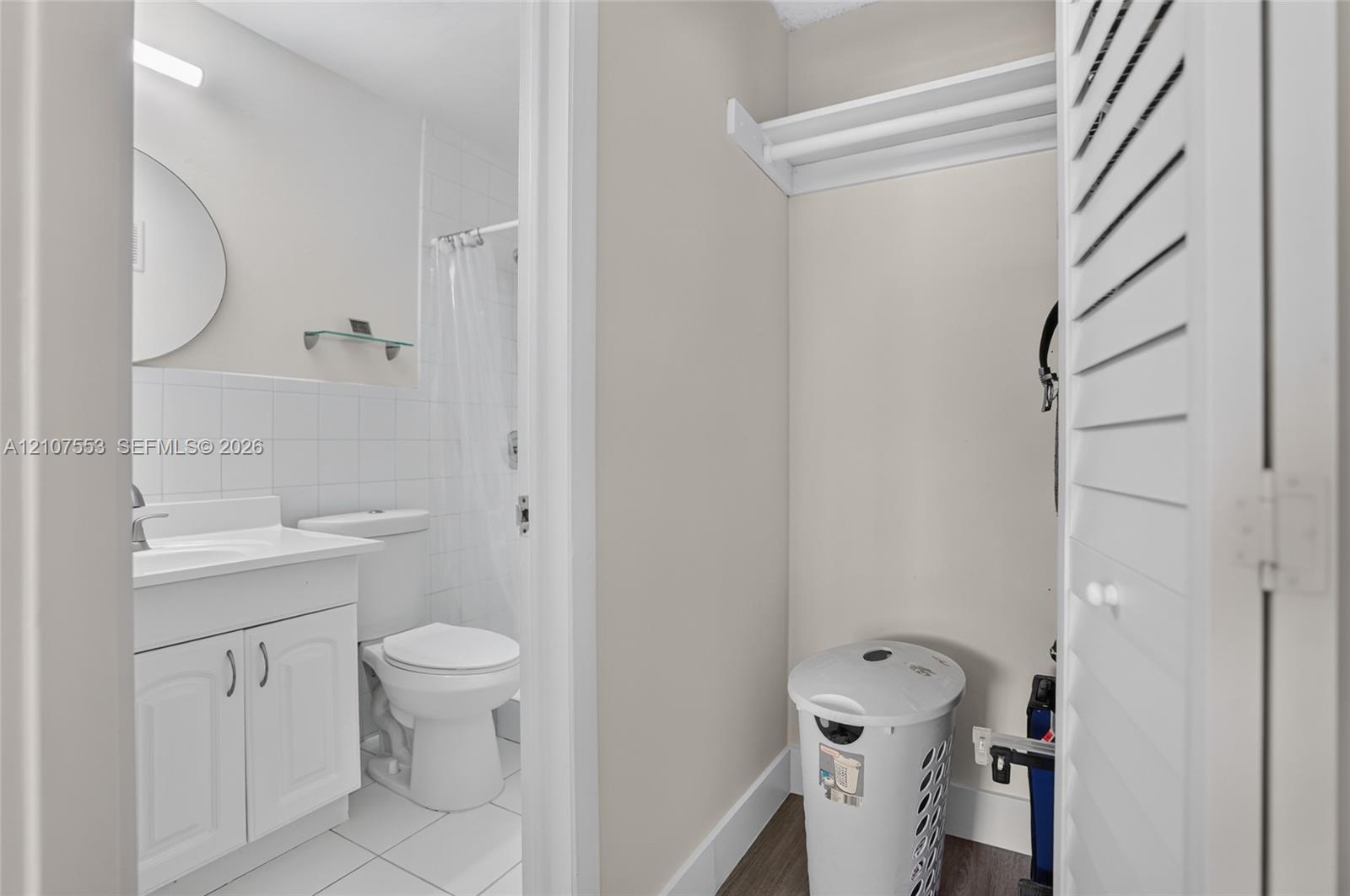 Bathroom featuring a white vanity cabinet, tiled shower surround, wall-mounted circular mirror, and louvered closet doors with integrated shelving