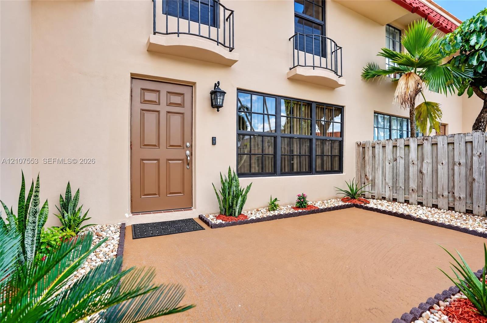 Stucco exterior featuring dual upper-level balconies with metal railings, a multi-pane grid window, and a paved front patio with decorative stone borders