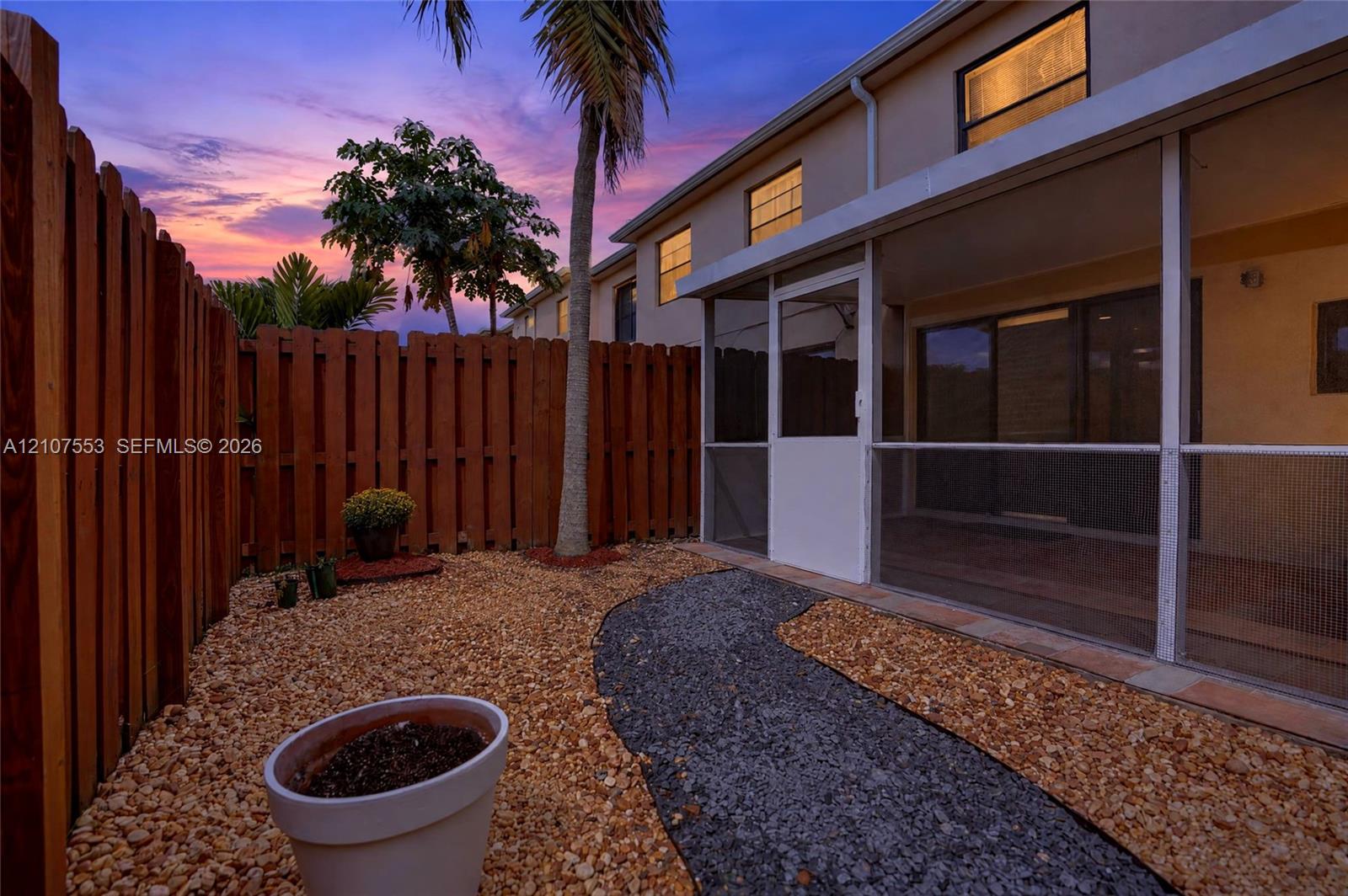 Screened-in patio with concrete flooring, stone-grounded yard, wood privacy fencing, and palm tree