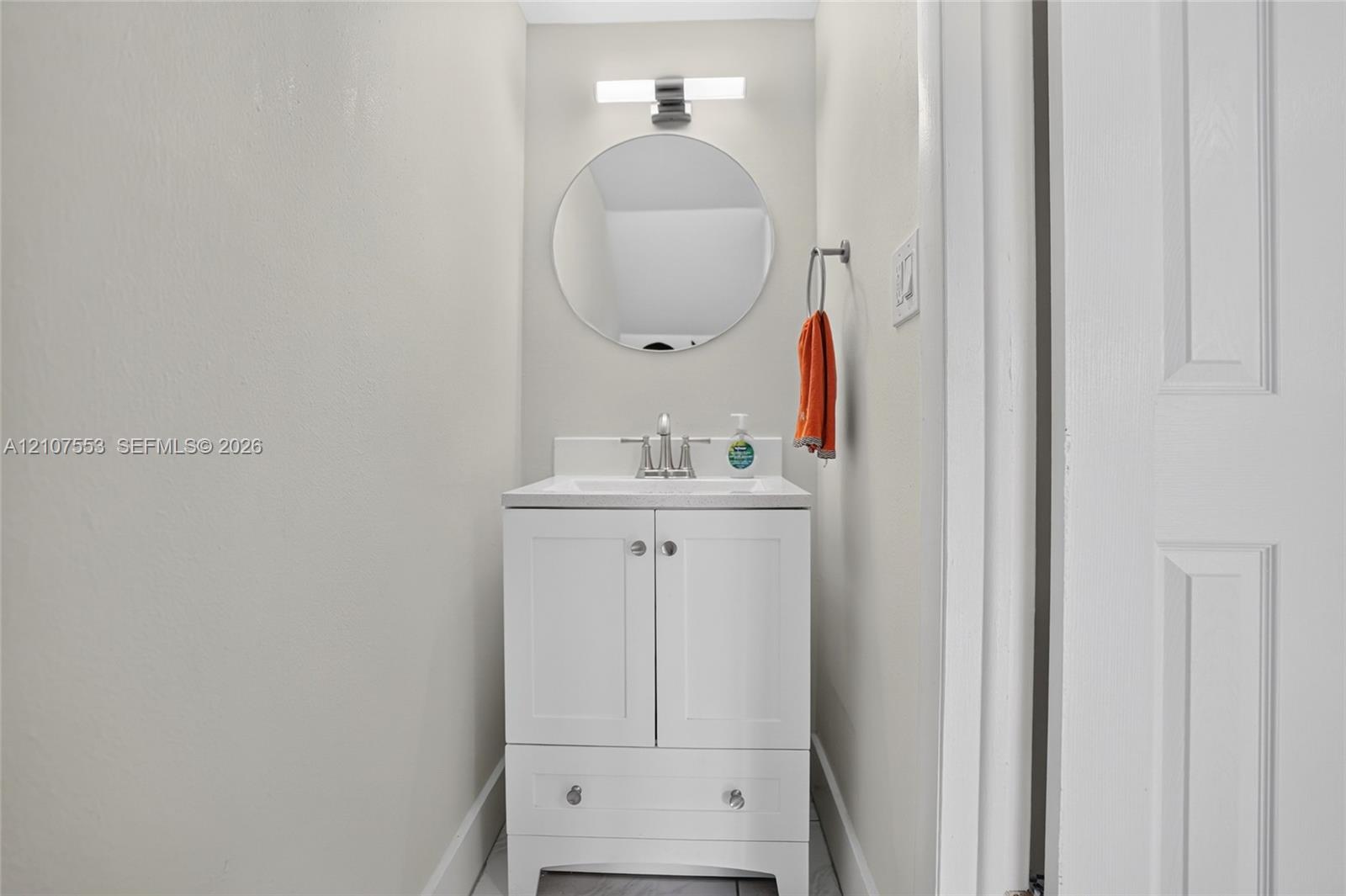 White vanity with lower storage drawer, integrated basin, brushed nickel hardware, circular wall-mounted mirror, and horizontal LED light fixture