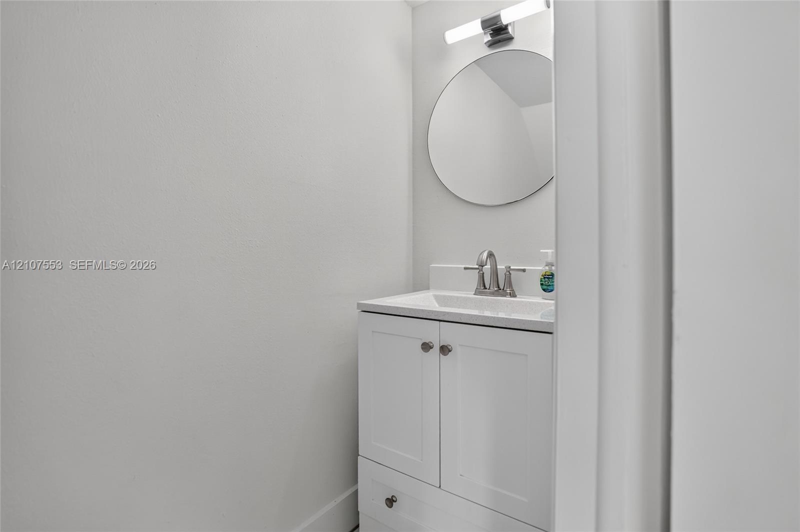 Shaker-style white vanity with a rectangular sink, satin-nickel faucet, circular wall mirror, and modern linear light fixture