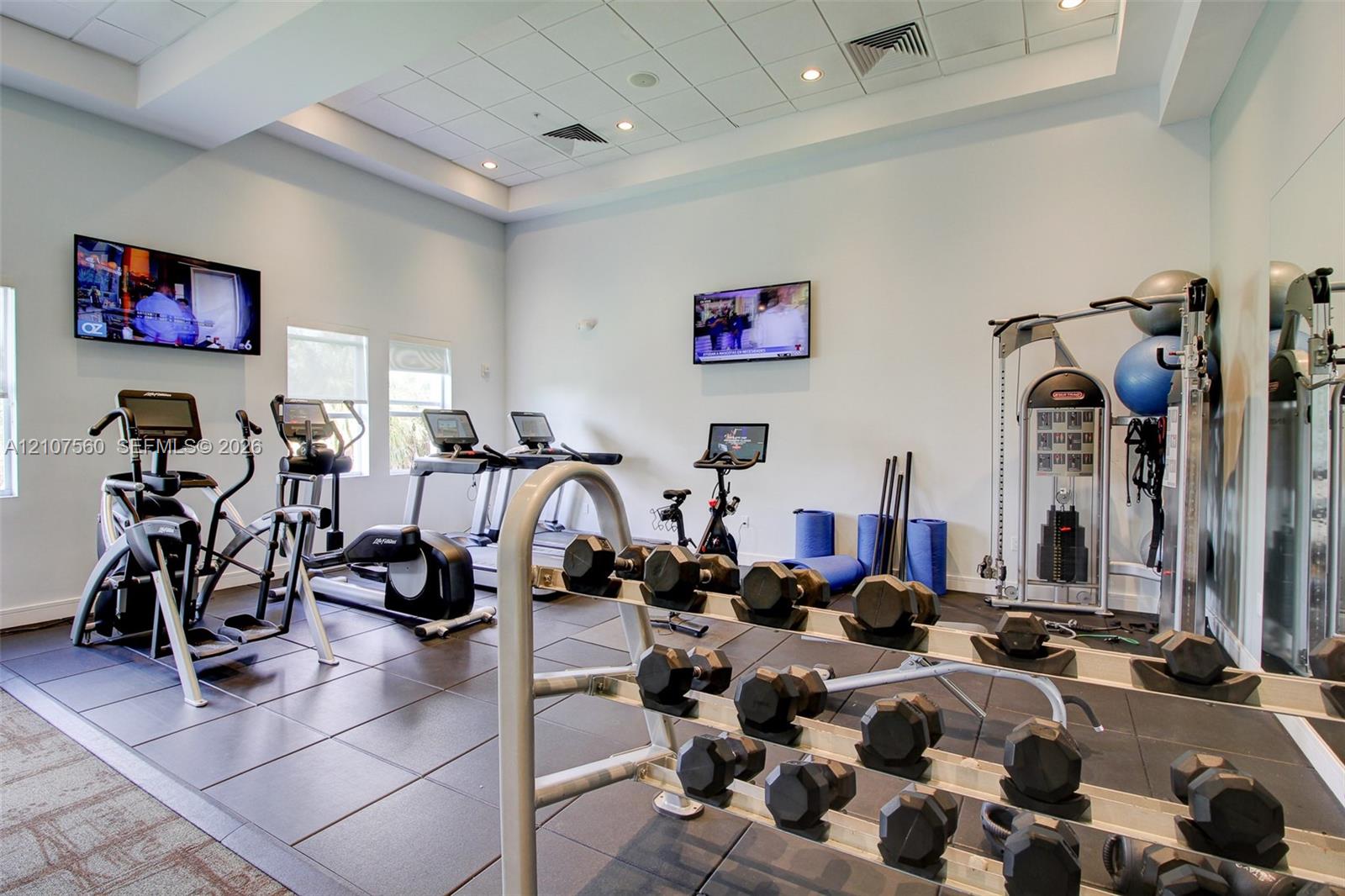 Fitness center featuring recessed tray ceilings, large-format tile flooring, wall-mounted televisions, and mirrored wall surfaces
