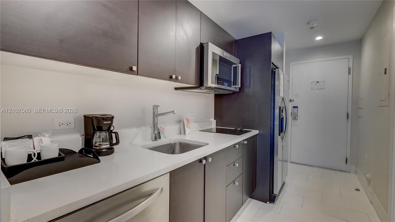 Kitchenette featuring flat-panel cabinetry, a solid-surface countertop, stainless steel sink with a high-arch faucet, a built-in microwave, and an integrated cooktop
