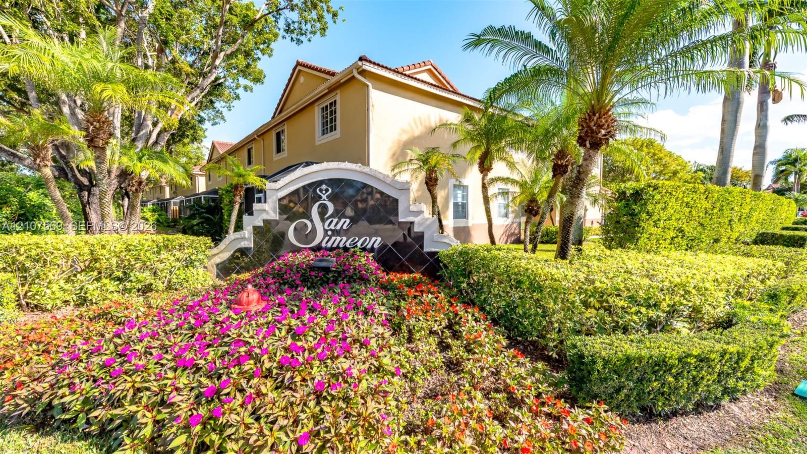 Community entrance featuring professional landscaping with manicured hedges, mature palm trees, floral beds, and custom masonry signage