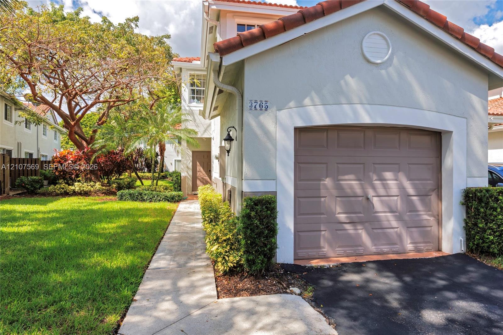Exterior facade featuring stucco siding, a tiled roof, a single-car garage, and a concrete walkway bordered by manicured landscaping