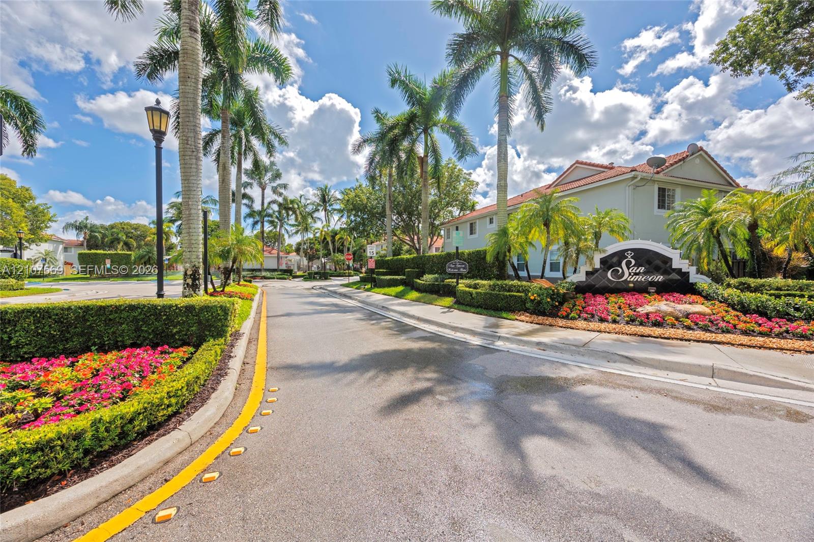 Paved entryway featuring mature palm trees, manicured hedges, colorful floral landscaping, and dedicated post-mounted street lighting