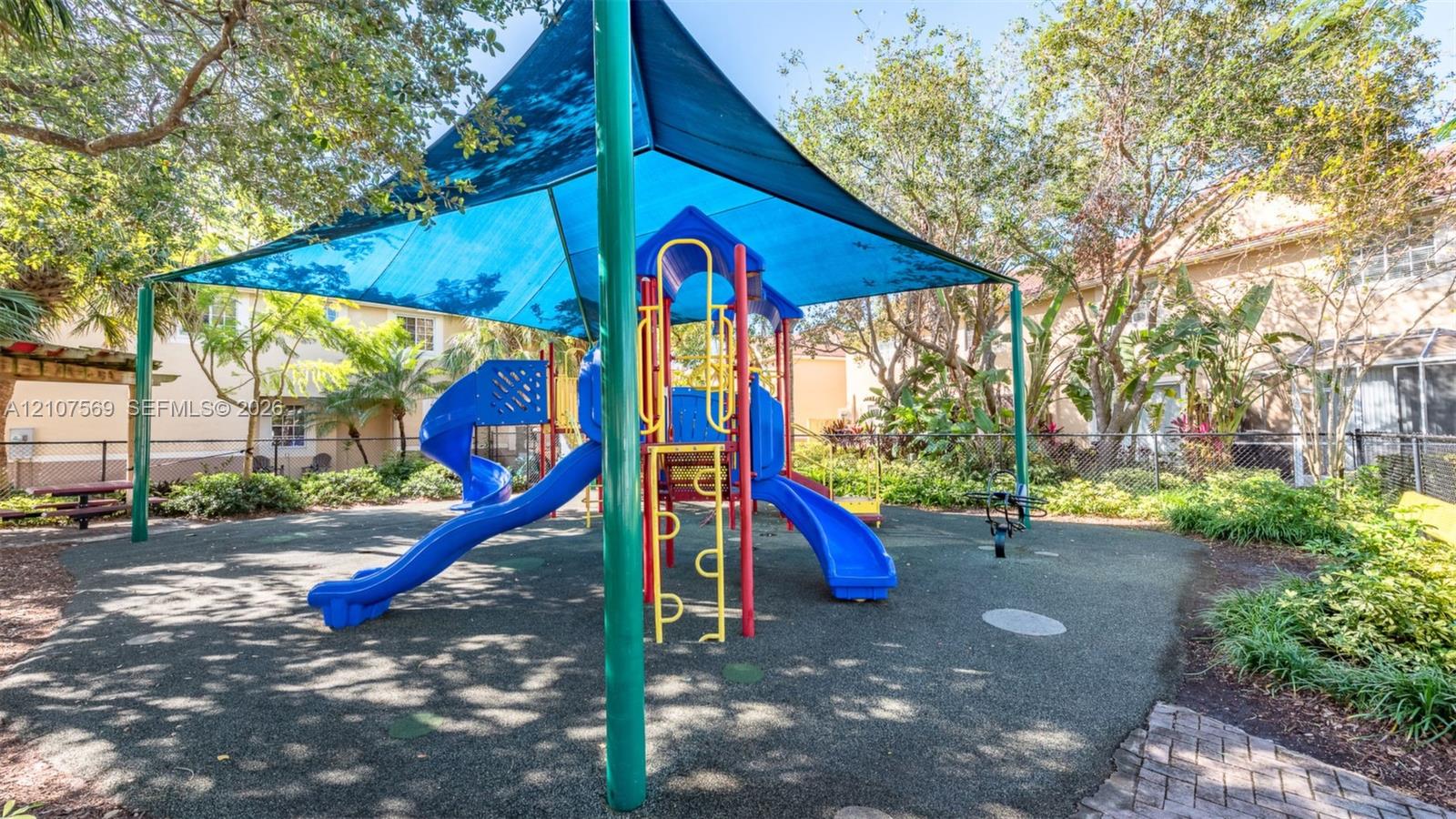 Playground featuring a central climbing structure with dual slides, a fabric shade canopy, rubberized flooring, and surrounding landscaping