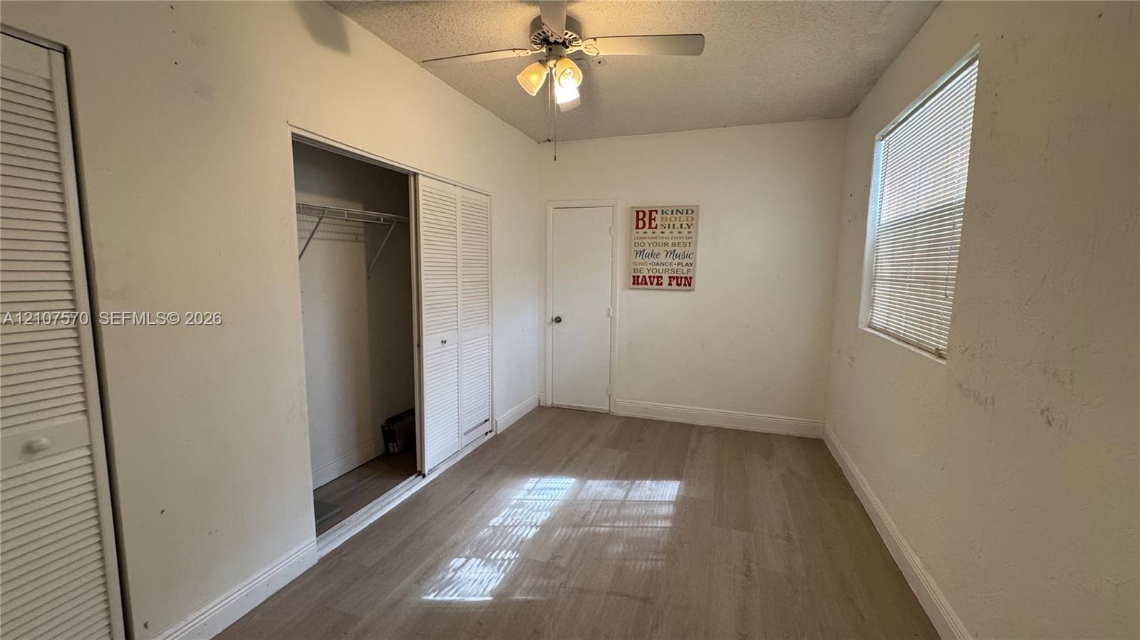 Room featuring wood-finish flooring, a ceiling fan with integrated lighting, two sets of louvered closet doors, and a window with horizontal blinds