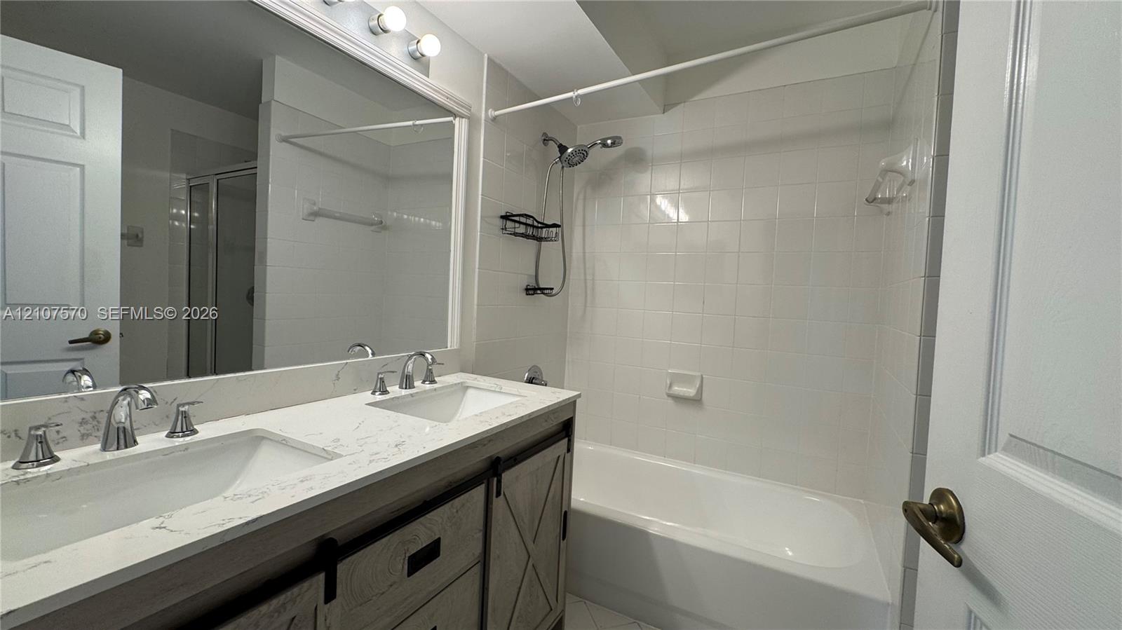 Dual-sink vanity with light-toned stone-patterned countertop, extensive wall-mounted mirror, white tiled shower-tub combination, and industrial-style sliding cabinet doors