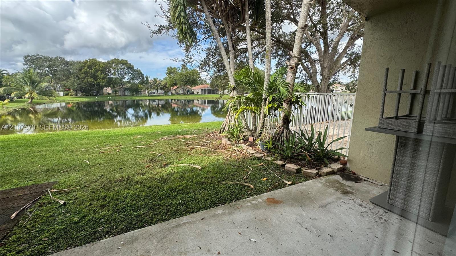 Concrete patio overlooking a pond, featuring mature palm trees, landscaped garden beds with brick borders, and a metal perimeter fence